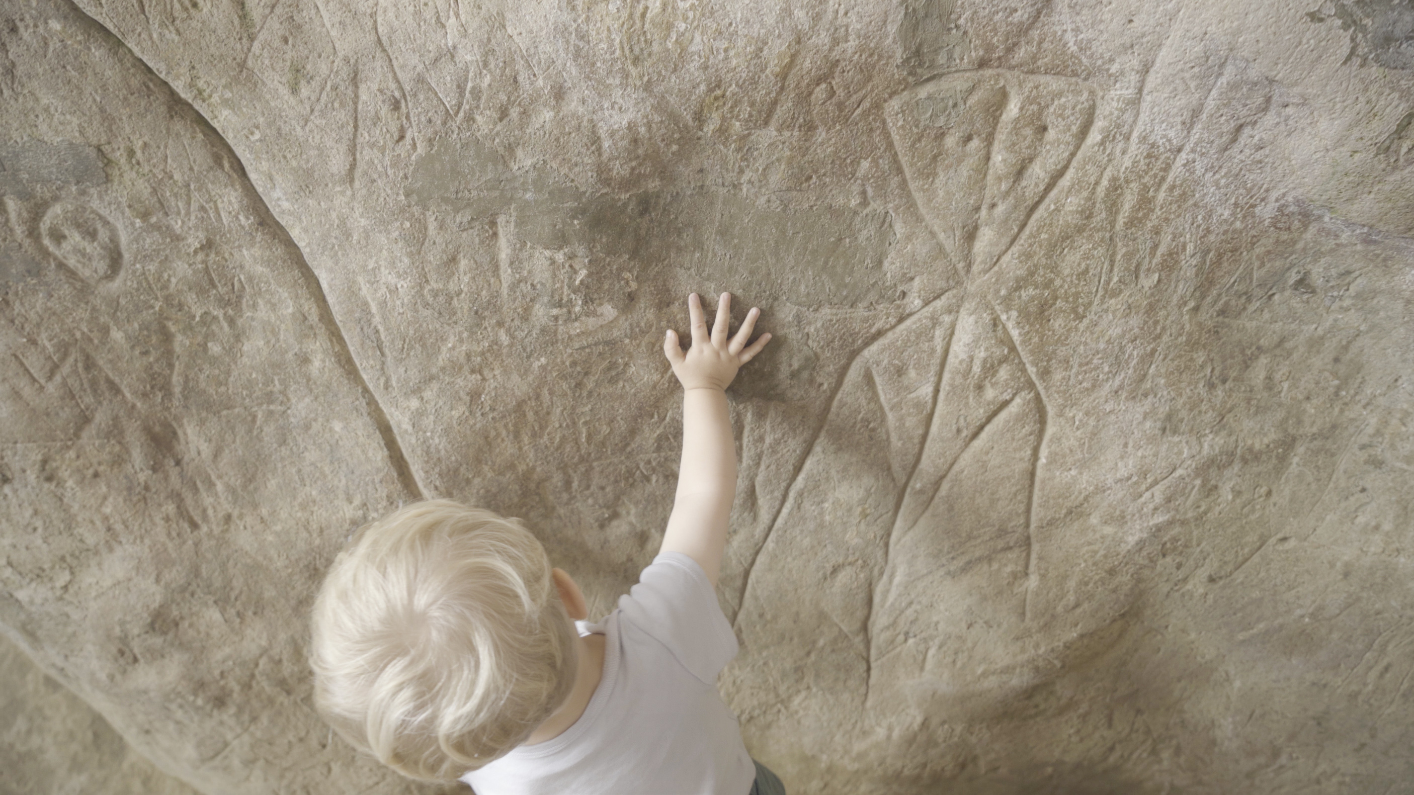 zicht op het blonde hoofd van een jonge kind dat de hand legt op een grote steen met daarin prehistorische tekens gegraveerd